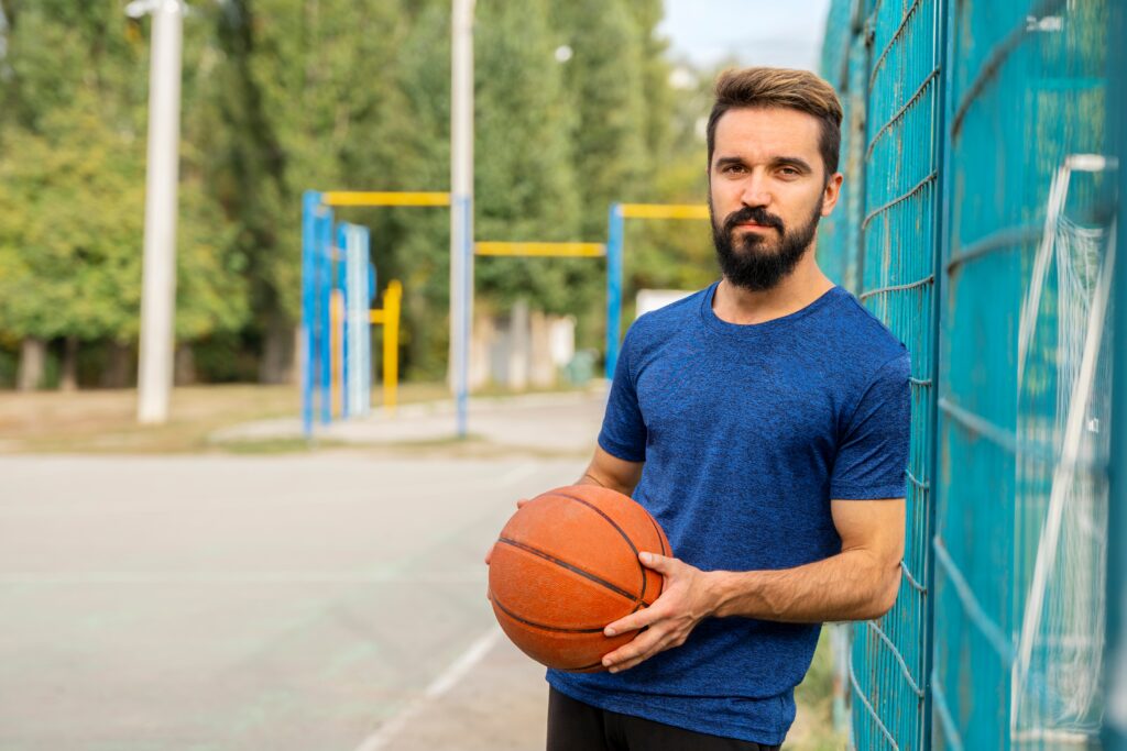 Brunette man with a beard leans against a blue fence outdoors while holding a basketball.
